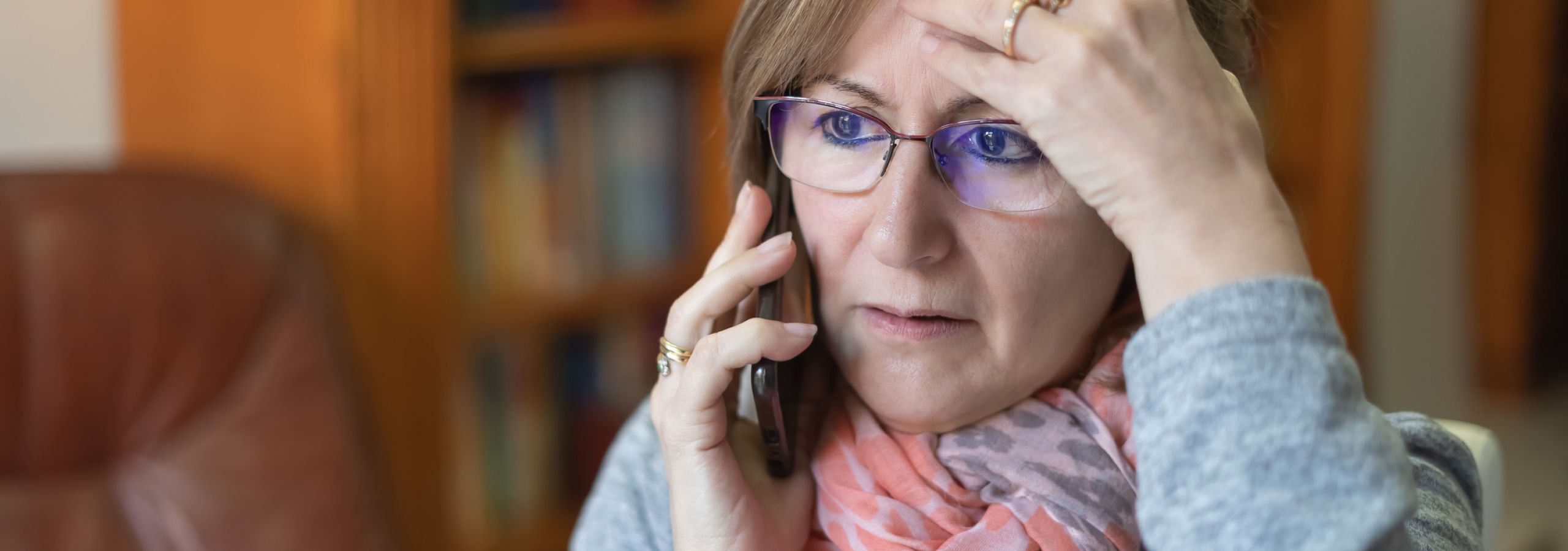 A middle-aged woman with glasses and a pink scarf answers a confusing scam call at home.