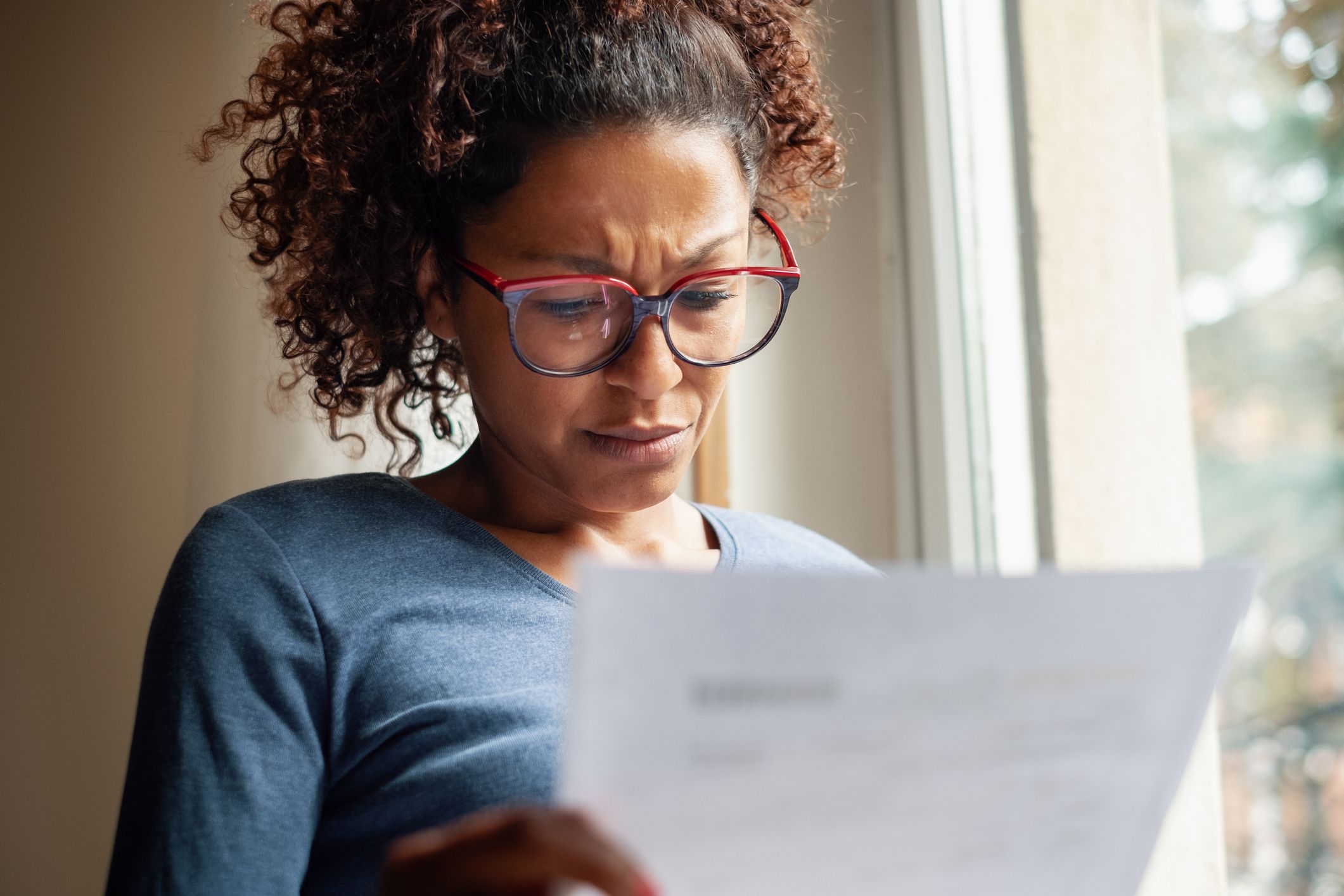 A black woman in red-framed reading glasses stands by a bright window and frowns at a bill containing fraudulent charges.