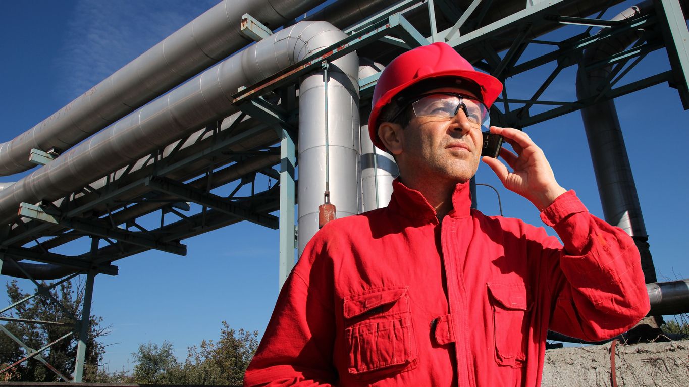 A man in a bright red jumpsuit, hard hat, and safety goggles stands outdoors in front of large refinery pipes to place a call.