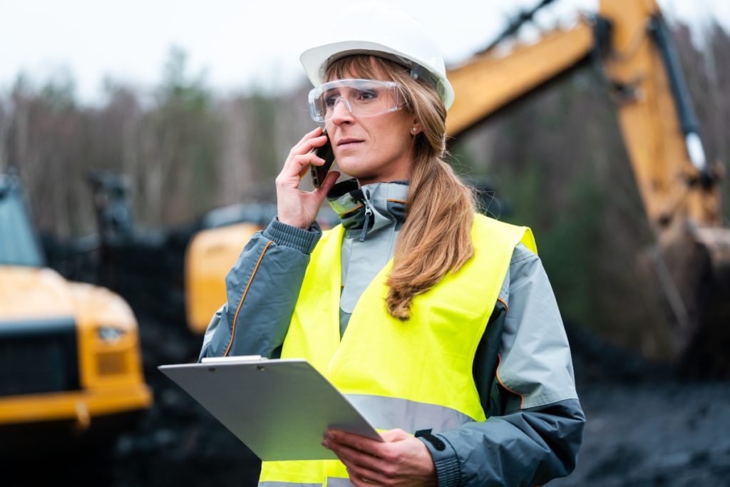 A construction forewoman in a white hard hat and holding a clipboard places a concerned call to a machine accident attorney.