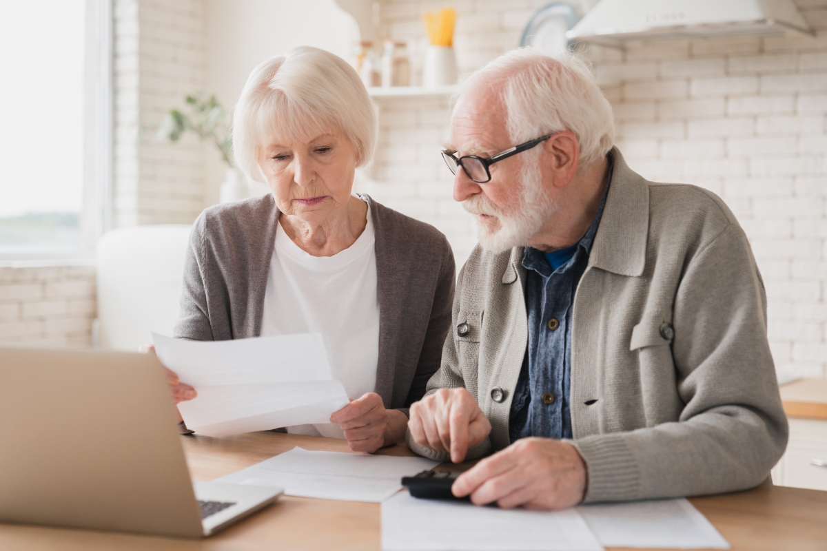 A white-haired couple calculates their bills and notices worrying overdraft fees in their finances.