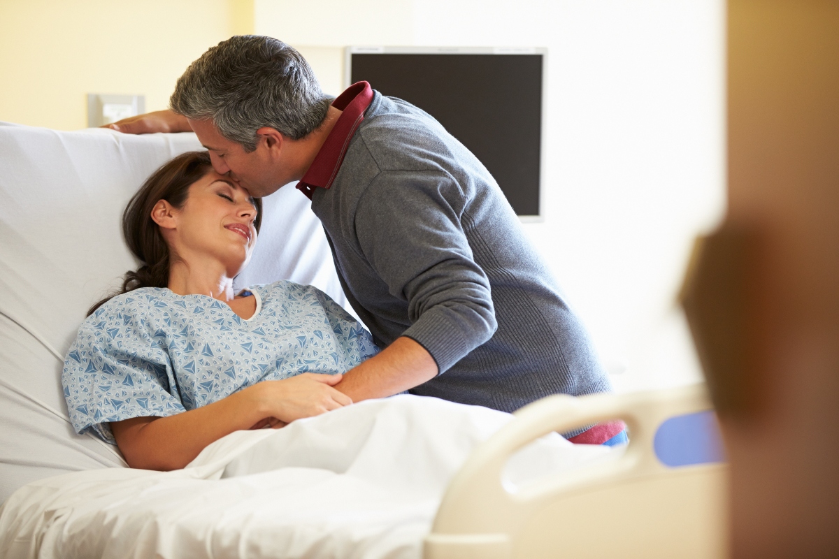 A woman is comforted in her hospital bed by her husband who leans down and kisses her forehead.