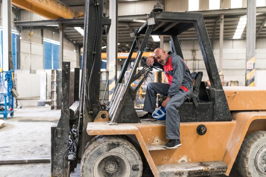  tired worker wipes sweat from his brow while sitting on a warehouse loader machine.